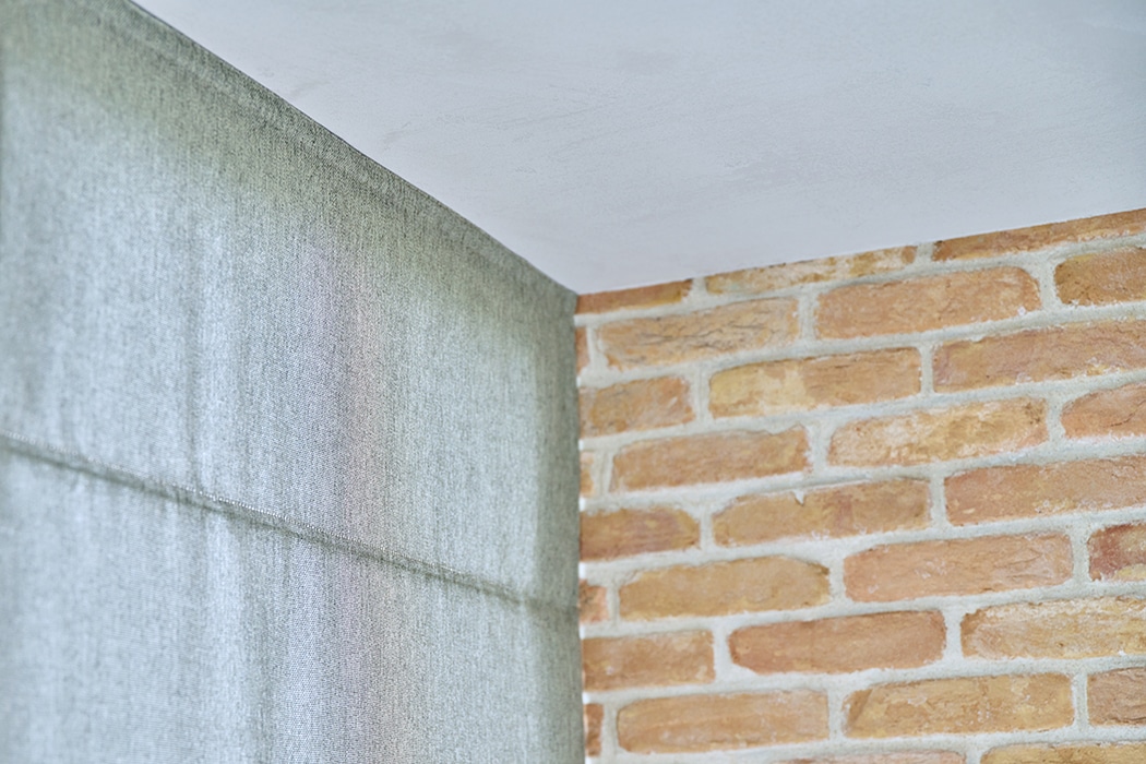 A corner of a room showing a brick wall, a section of ceiling, and a light-coloured fabric curtain.