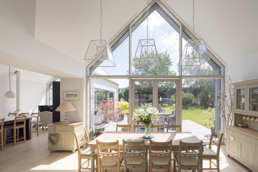 A bright dining area with large triangular windows overlooks a garden. A wooden table with chairs sits in the centre, surrounded by light-coloured furniture and modern pendant lights.