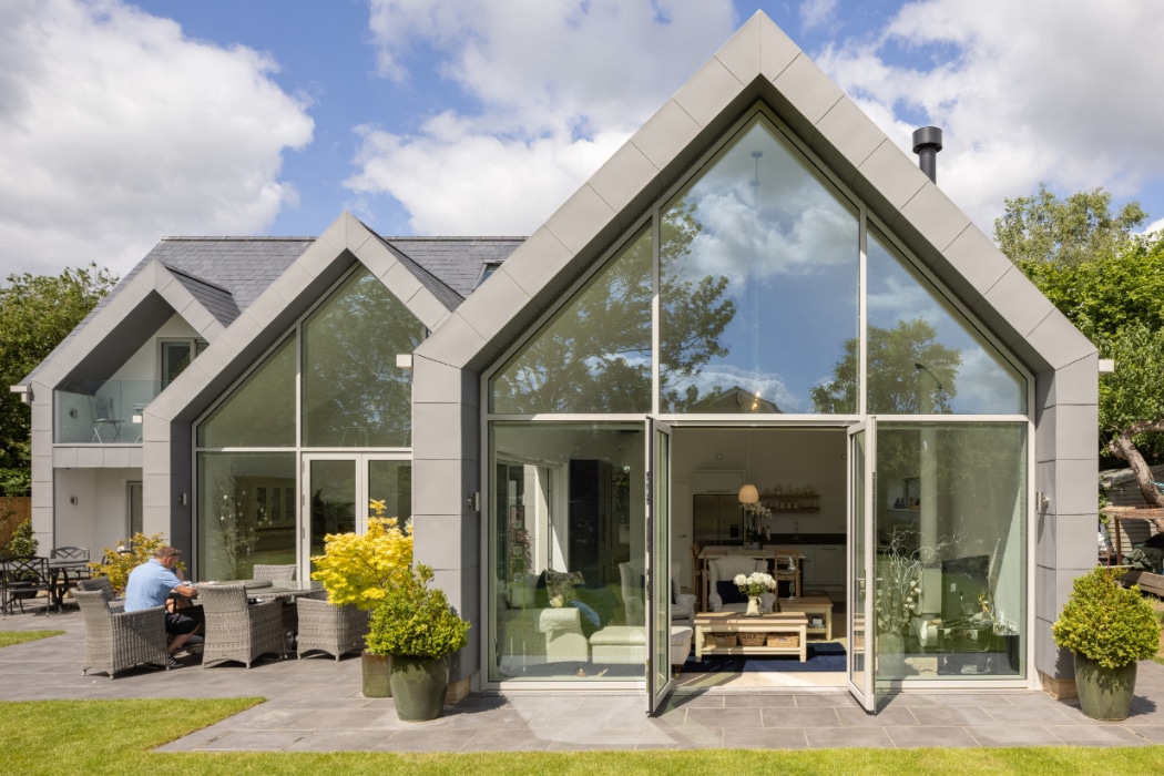 Modern two-storey house with large glass windows and doors, outdoor seating area, and landscaped garden on a sunny day.