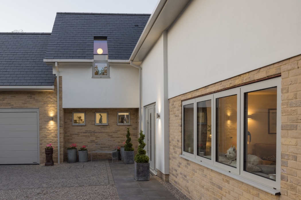 Modern house exterior with light brick and white walls, grey roof tiles, large front windows, and potted plants by the entrance. A white dog is visible through the window.