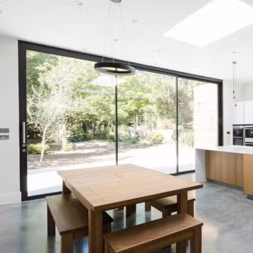 Modern kitchen and dining area with wooden table and benches, expansive glass sliding door, and a scenic view of the green garden outside.