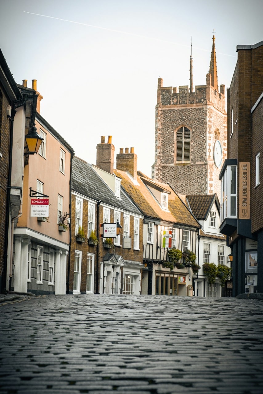 A cobbled street lined with historic buildings leads to a stone church with a tall clock tower in the background.
