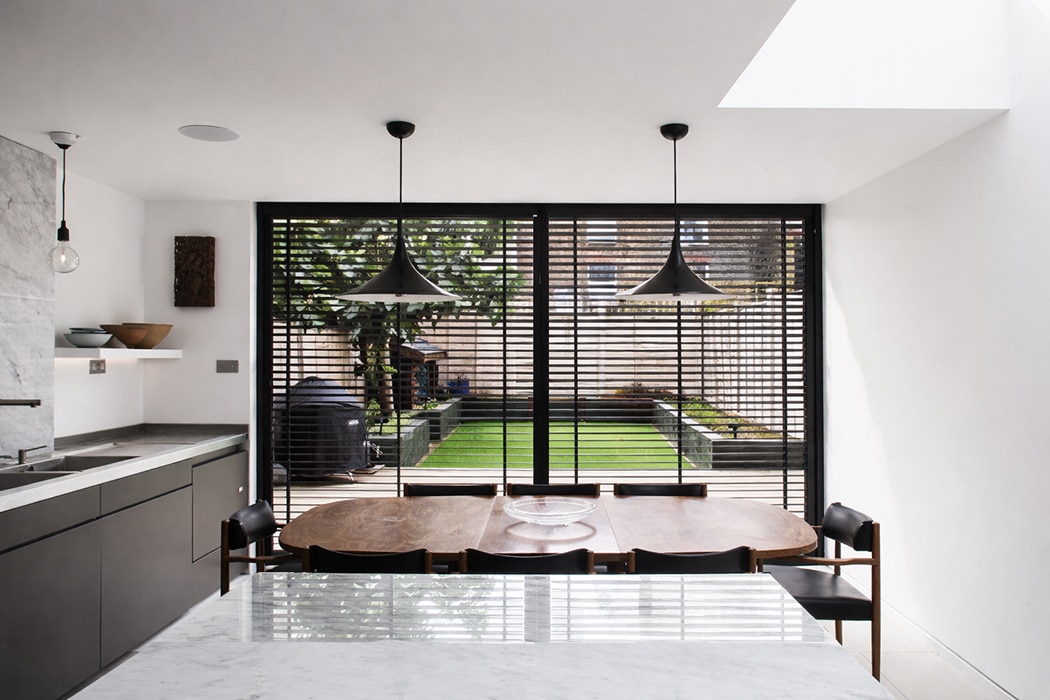 Modern kitchen and dining area with marble worktop, wooden table, black chairs, and large windows with blinds overlooking a landscaped back garden.