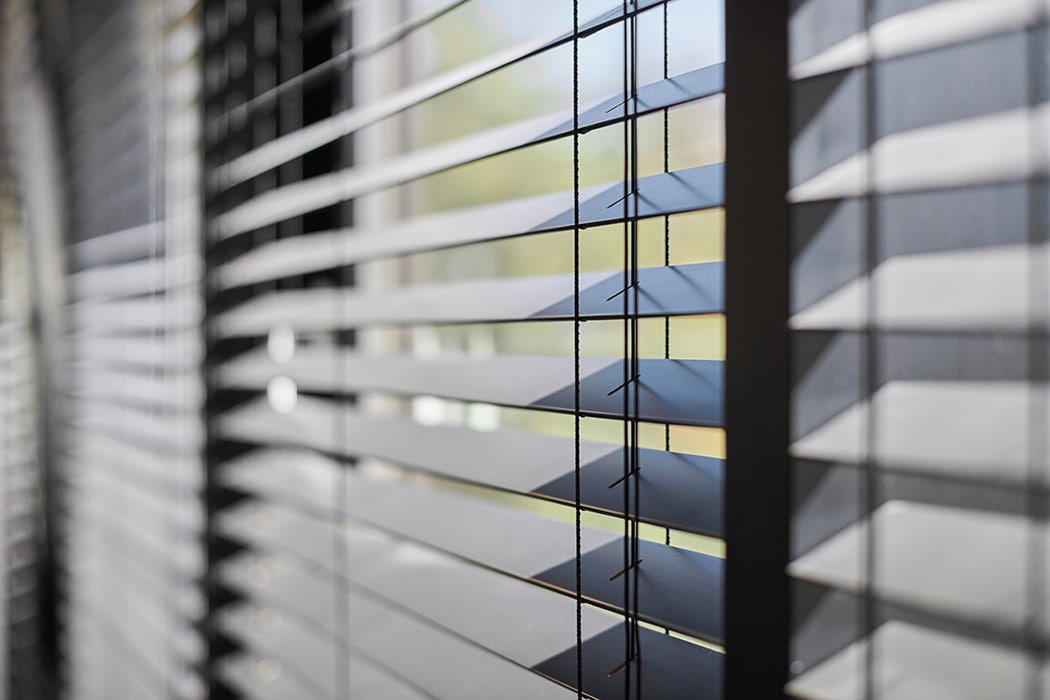 Close-up view of partially open horizontal window blinds with light filtering through, creating lines and shadows on the slats.