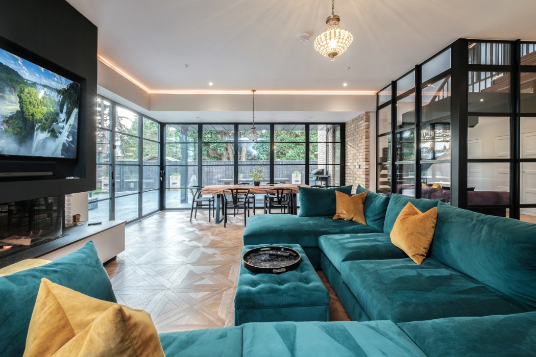 Modern open-plan living room with teal velvet seating, featuring slim-profile aluminium bifold doors opening to a garden terrace and dining area beyond.