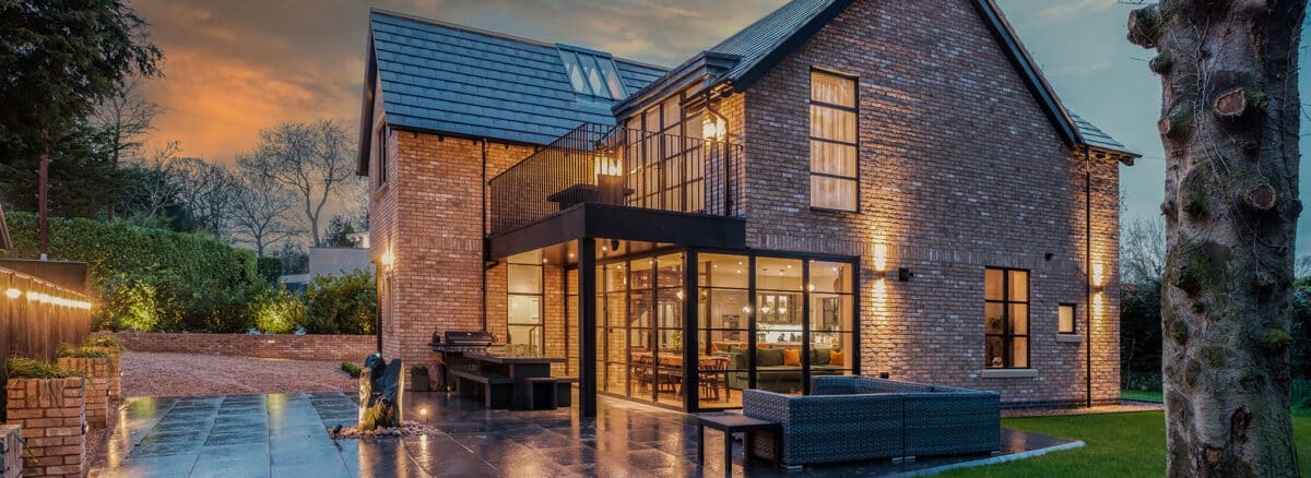 Modern two-storey brick house with large glass windows and patio seating, illuminated at dusk with outdoor lighting.