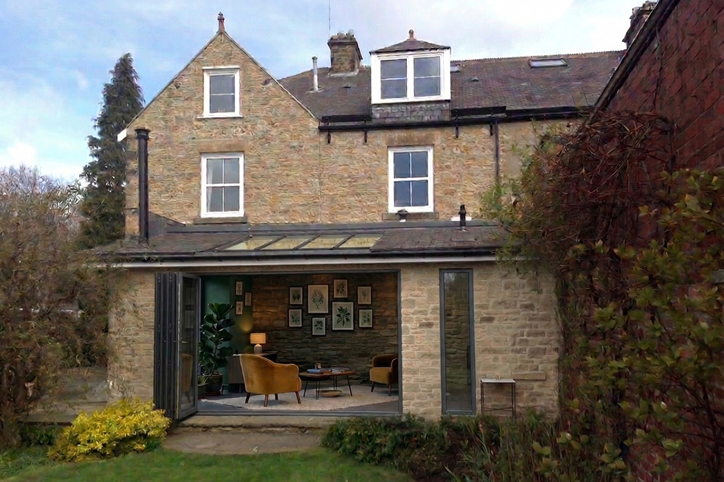 A stone house with large glass doors and structural glass roofs opens to a garden. Inside, a modern living area with armchairs, plants, and framed artwork is visible.