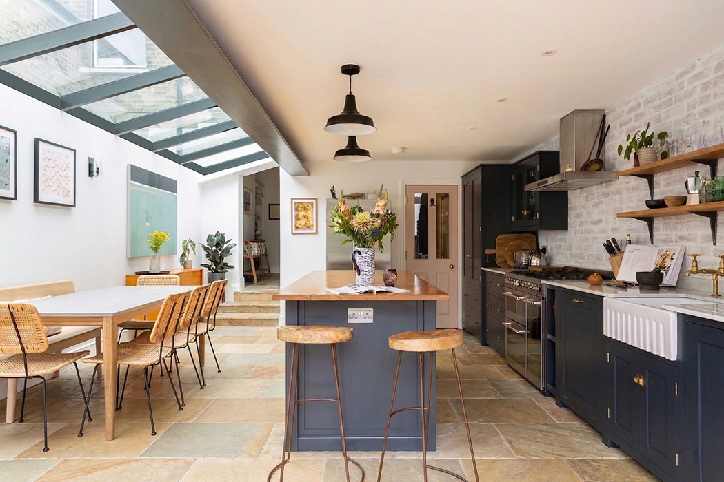 Modern kitchen with navy cupboards, a central island with barstools, dining table with chairs, and a skylight ceiling featuring structural glass roofs, plus decor including plants and artwork on the walls.