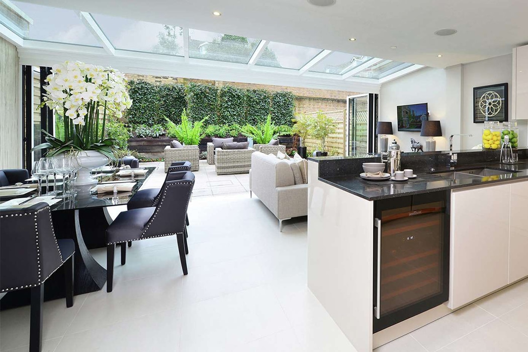 Modern open-plan kitchen and dining area leads to a patio with outdoor seating; structural glass roofs and folding doors provide natural light, with greenery visible outside.