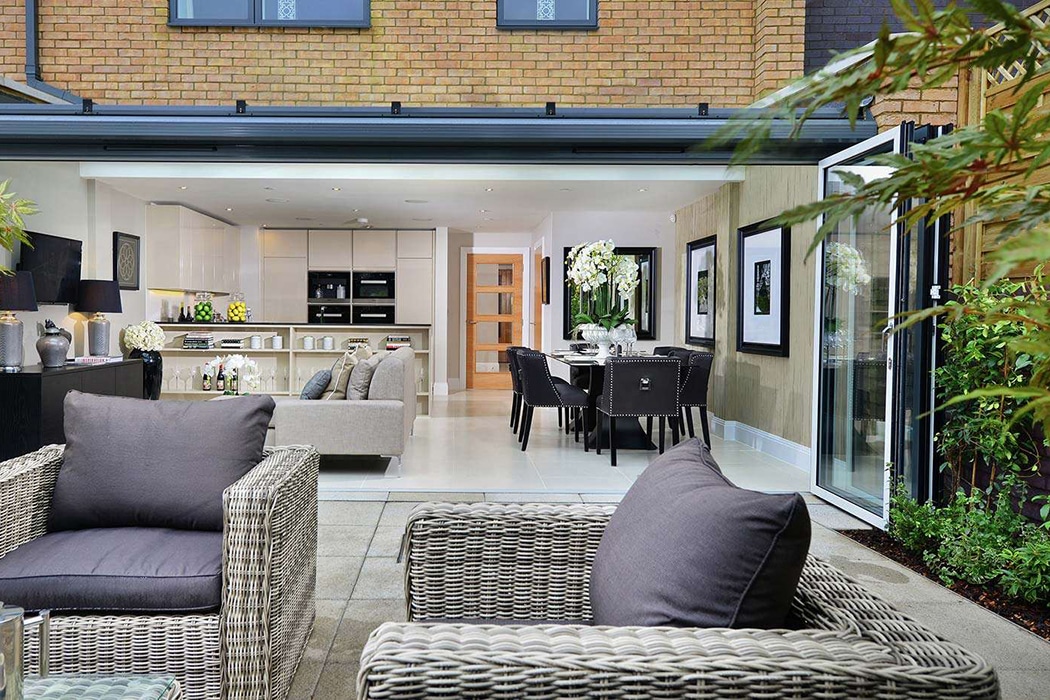 Modern open-plan kitchen, dining, and living area seen from a patio with wicker chairs and cushions; folding glass doors and structural glass roofs seamlessly connect the indoor and outdoor spaces.