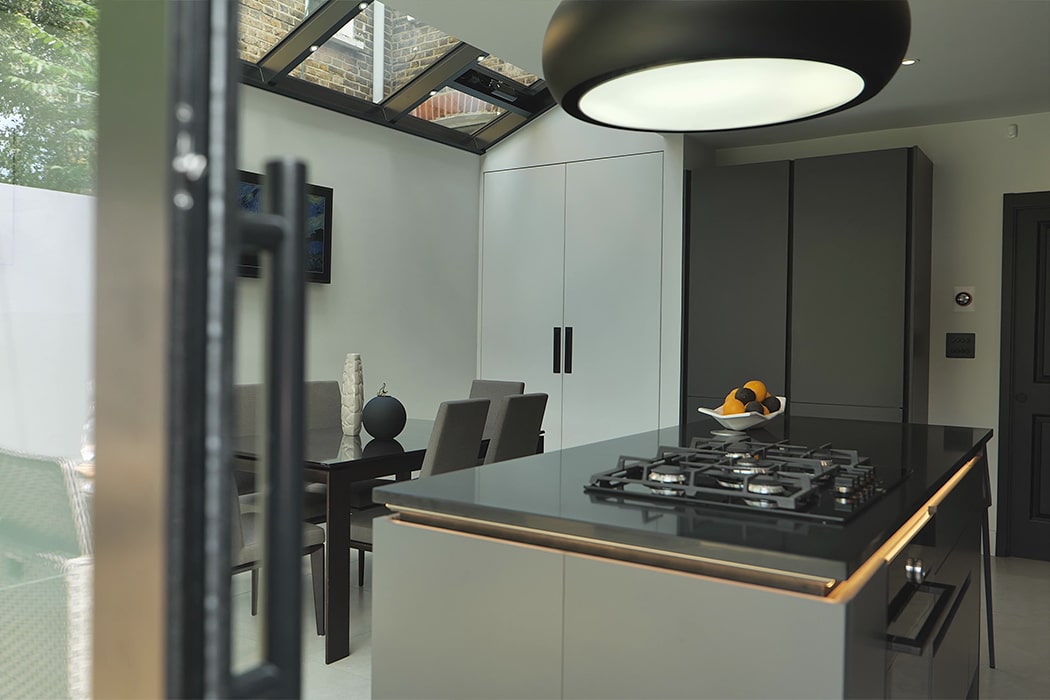 Modern kitchen with a black island worktop, built-in gas hob, bowl of fruit, dining table with chairs, large overhead light, and skylight windows enhanced by structural glass roofs letting in natural light.