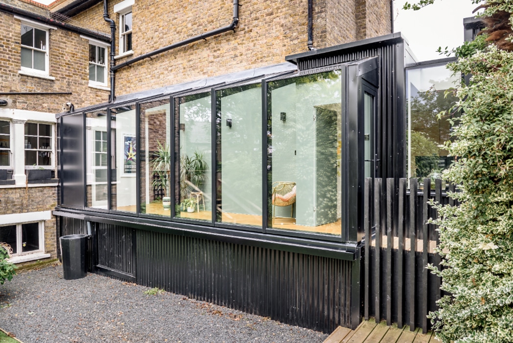 A modern glass and metal extension with floor-to-ceiling windows and sleek structural glass roofs is attached to a traditional brick house, overlooking a gravel path and garden.