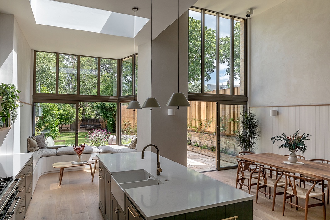 Contemporary kitchen with double-height glazing overlooking garden, featuring pendant lights and wooden dining table.