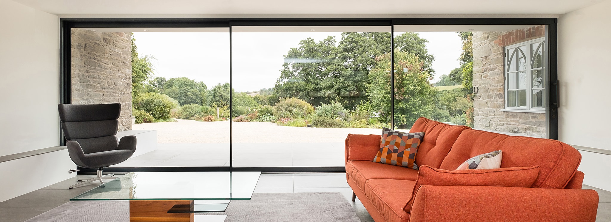 Modern living space with orange sofa and black chair overlooking countryside through expansive sliding glass doors and windows.