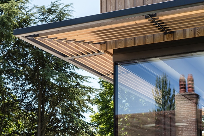 Modern timber-framed pergola with slatted roof design overlooking brick residential property with tall chimneys.