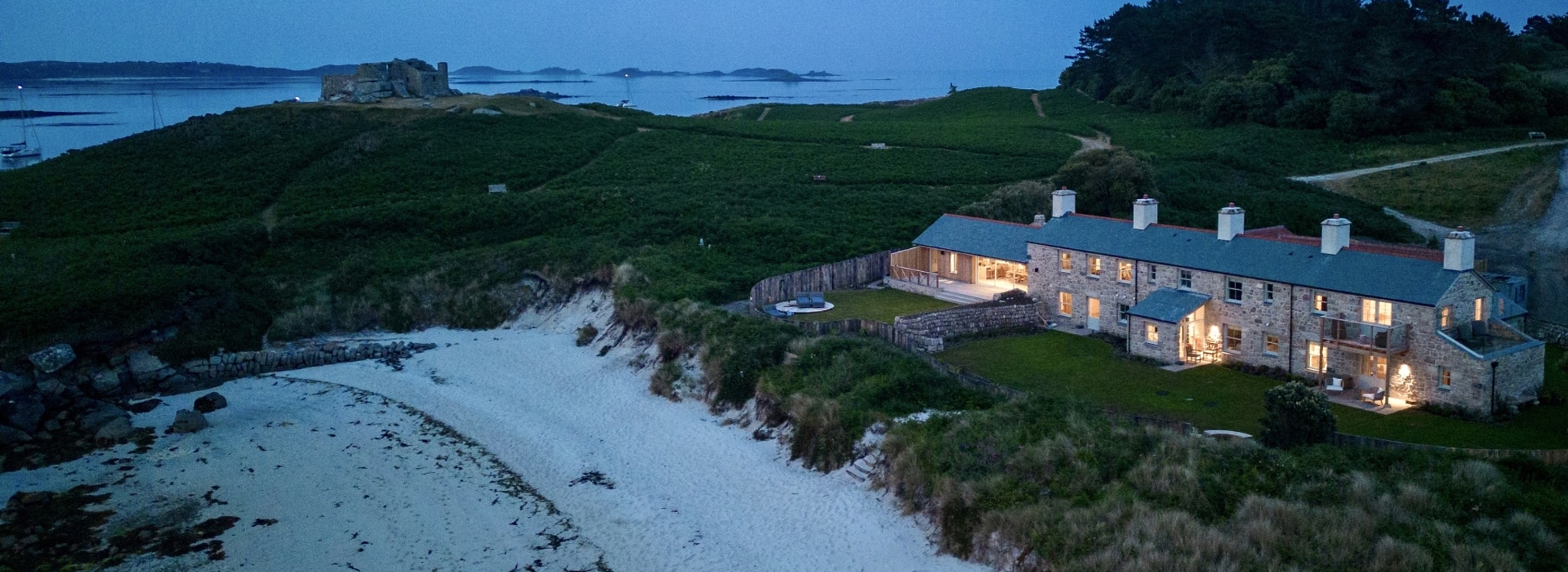 Blockhouse Cottage on Tresco, one of the Isles of Scilly, with illuminated windows overlooking beach at dusk, featuring extensive glazing and sea views.